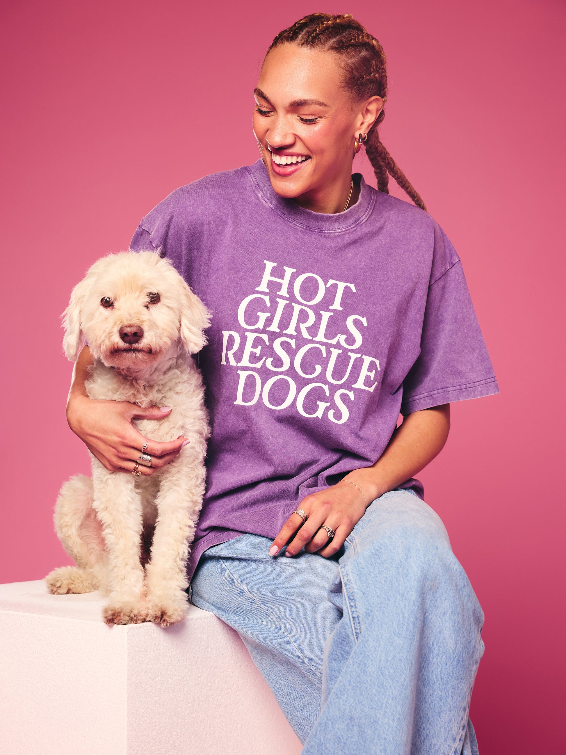 Person wearing a purple 'Hot Girls Rescue Dogs' t-shirt holding a white dog against a pink background