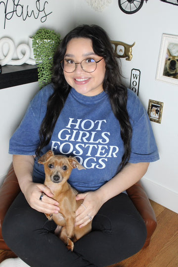 Woman wearing a blue t-shirt with 'Hot Girls Foster Dogs' text, holding a small dog indoors.