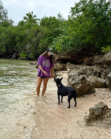 Woman in a purple T-shirt with text  Hot Girl Rescue  Dogs and a cap, standing on a beach with a black dog.