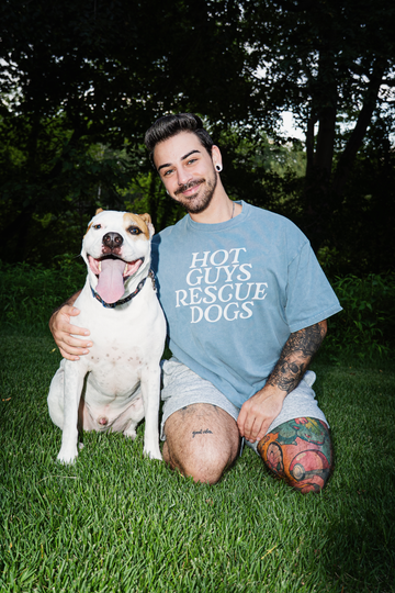 Man wearing a 'Hot Guys Rescue Dogs' t-shirt is sitting on the grass with a dog.
