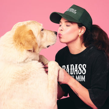 Woman wearing a 'BADASS MOM' shirt and green cap with a dog on a pink background