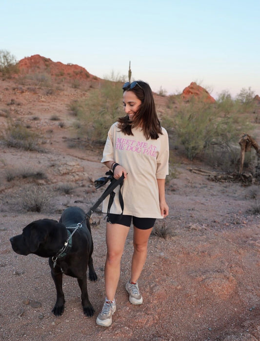 A woman walking a black dog and wearing a T-shirt with Meet Me At The Dog Park text 