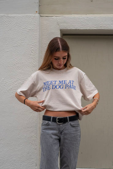 Person wearing a white t-shirt with blue Meet Me at the Dog Park text standing against a plain wall.