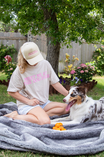 A person sitting on a blanket with a dog and wearing a T-shirt with  Meet Me at the Dog Park text.