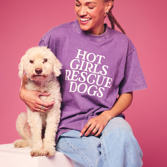 Person wearing a purple 'Hot Girls Rescue Dogs' t-shirt holding a white dog against a pink background